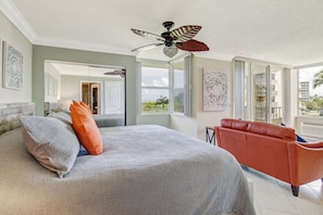 A bedroom with a bed, a red sofa, a ceiling fan, and large windows. - Estero Beach & Tennis 207 C (Fort Myers Beach)