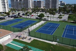A tennis court complex with multiple courts, surrounded by a parking area and a row of high-rise buildings. - Estero Beach & Tennis 207 C (Fort Myers Beach)