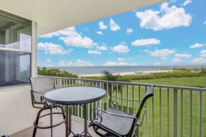 A balcony with a table and chairs, overlooking a beach and ocean. - Estero Beach & Tennis 207 C (Fort Myers Beach)