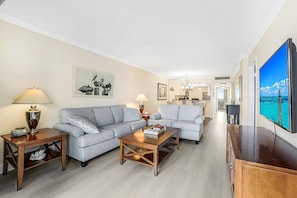 Ein geräumiges Wohnzimmer mit grauem Sofa, einem Couchtisch und einem Fernseher auf einer Holzkommode. An der Wand hängt ein Gemälde und im Hintergrund ist ein Essbereich zu sehen.