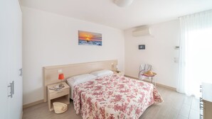 Una habitación de hotel con cama, mesita de noche, lámpara, un cuadro de un atardecer, equipo de aire acondicionado y ventana con cortinas.