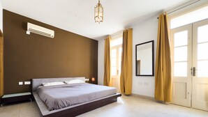 A bedroom with a bed, a bedside table, an air conditioning unit, a lamp, a mirror, and a door with curtains.