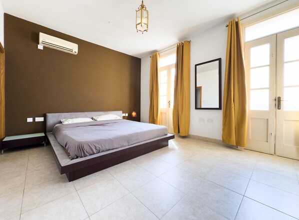 Een slaapkamer met een bed, nachtkastje, airconditioning, lamp, spiegel en een deur met gordijnen.