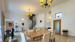A spacious dining area with a large wooden table and white chairs, a spiral staircase, and a chandelier.