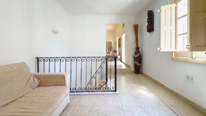 A hallway with a beige sofa, a decorative metal railing, a large vase, and a window with open shutters.