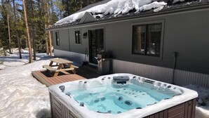 Un jacuzzi en un área exterior nevada con una terraza de madera y una casa al fondo.