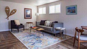 A modern living room with a grey sofa, two armchairs, a coffee table, and a patterned rug. There are two windows, a lamp, and wall art. - The Fireweed Gardens (Anchorage)