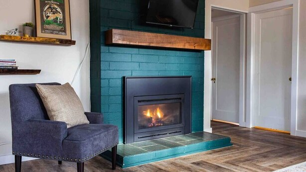 A living room with a teal brick fireplace, a dark blue armchair, and a mounted television. - The Fireweed Gardens (Anchorage)