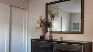 A bedroom with a dark dresser, a mirror with a decorative frame, and a vase with flowers. - The Fireweed Gardens (Anchorage)
