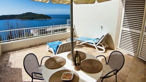 Un balcon avec une table dressée pour deux personnes, une bouteille et un bol de noix. On y trouve deux fauteuils et un grand parasol. La vue comprend une étendue d’eau et une île lointaine.