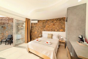 A modern hotel room with a stone wall, a bed with a white headboard, a glass door leading to a balcony, and a small bedside table with a lamp. - Canes Villa Heated Pool (Agios Nikolaos)