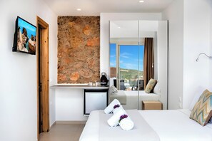 A hotel room with a stone wall, a bed with towels, a small kitchenette, and a view of the outdoors.