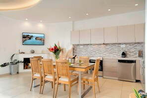 A modern kitchen with a dining area, featuring a flat-screen TV, wooden chairs, a vase with flowers, and stainless steel appliances.