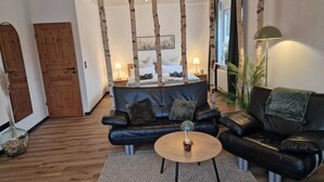 A living room with a black leather sofa, a round coffee table, and a wooden floor. There are two lamps on side tables, and a door is visible in the background. - Studio "ton Barkenboom" (Esens)