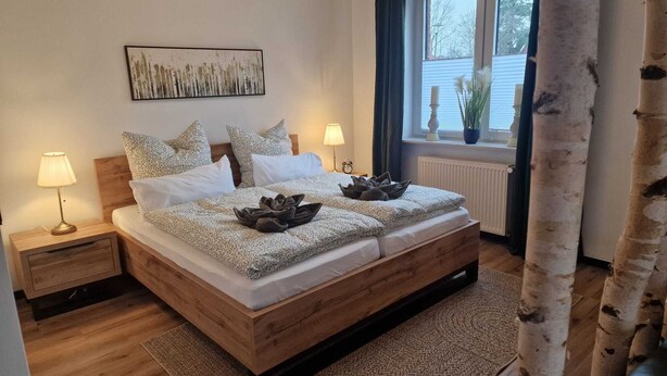 A bedroom with a wooden bed, two bedside tables with lamps, a window with a view, and a painting on the wall. - Studio "ton Barkenboom" (Esens)