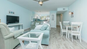 Een moderne woonkamer met een flatscreen televisie, een bank, een glazen salontafel en een eethoek met witte stoelen.