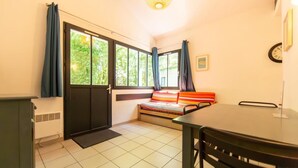 A small room with a bed, a table, and a chair. There is a window with curtains, a radiator, and a clock on the wall. - Maisonnette St Brice 44 for 5 persons (Arès)