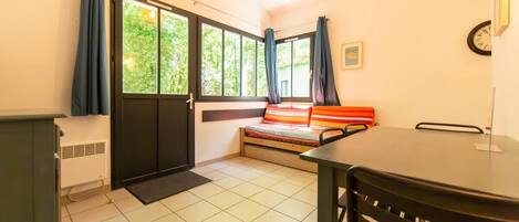 A small room with a bed, a table, and a chair. There is a window with curtains, a radiator, and a clock on the wall.