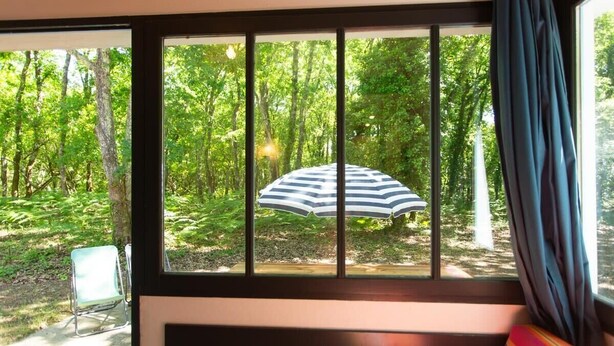 A room with a large window overlooking a forested area, a striped umbrella outside, and a chair by the window. - Maisonnette St Brice 44 for 5 persons (Arès)