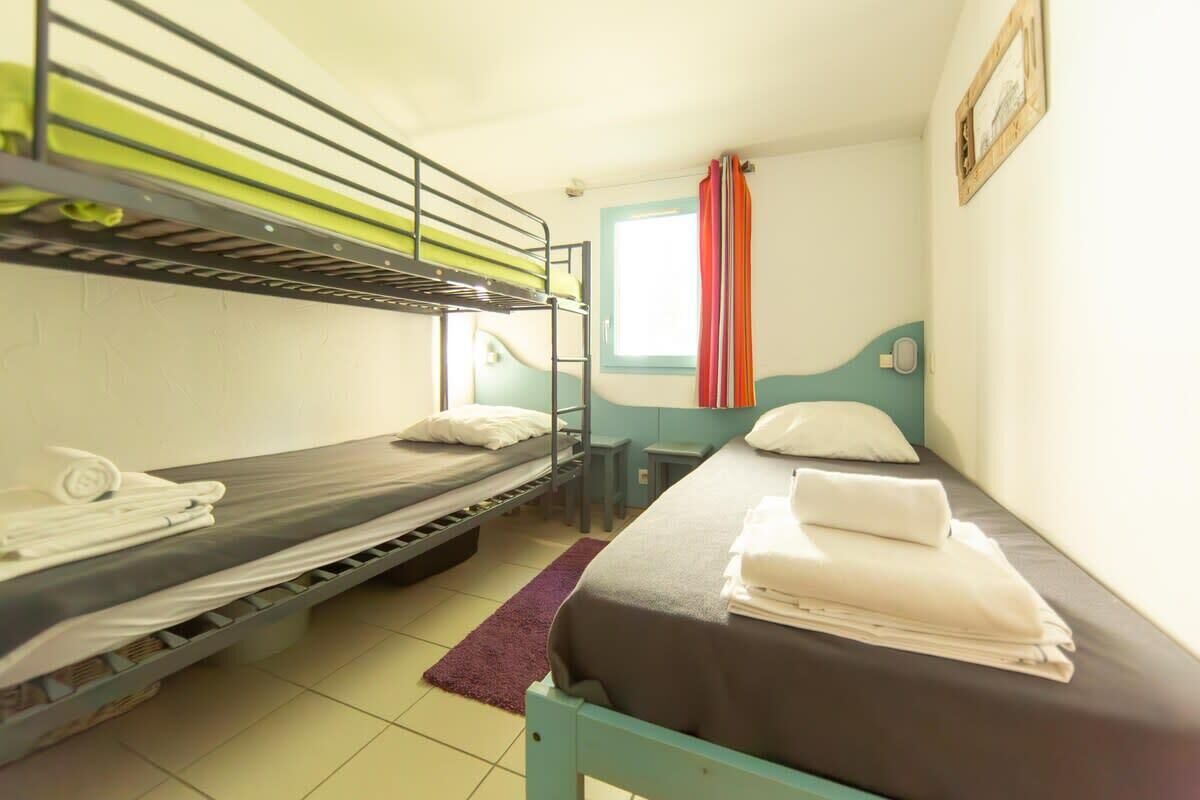 A dormitory room with bunk beds, a single bed, a window with curtains, and a small bedside table.