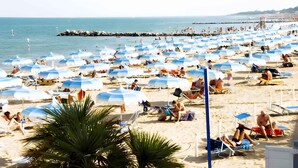 A beach with numerous umbrellas and sun loungers, a pier in the background, and a clear sky.