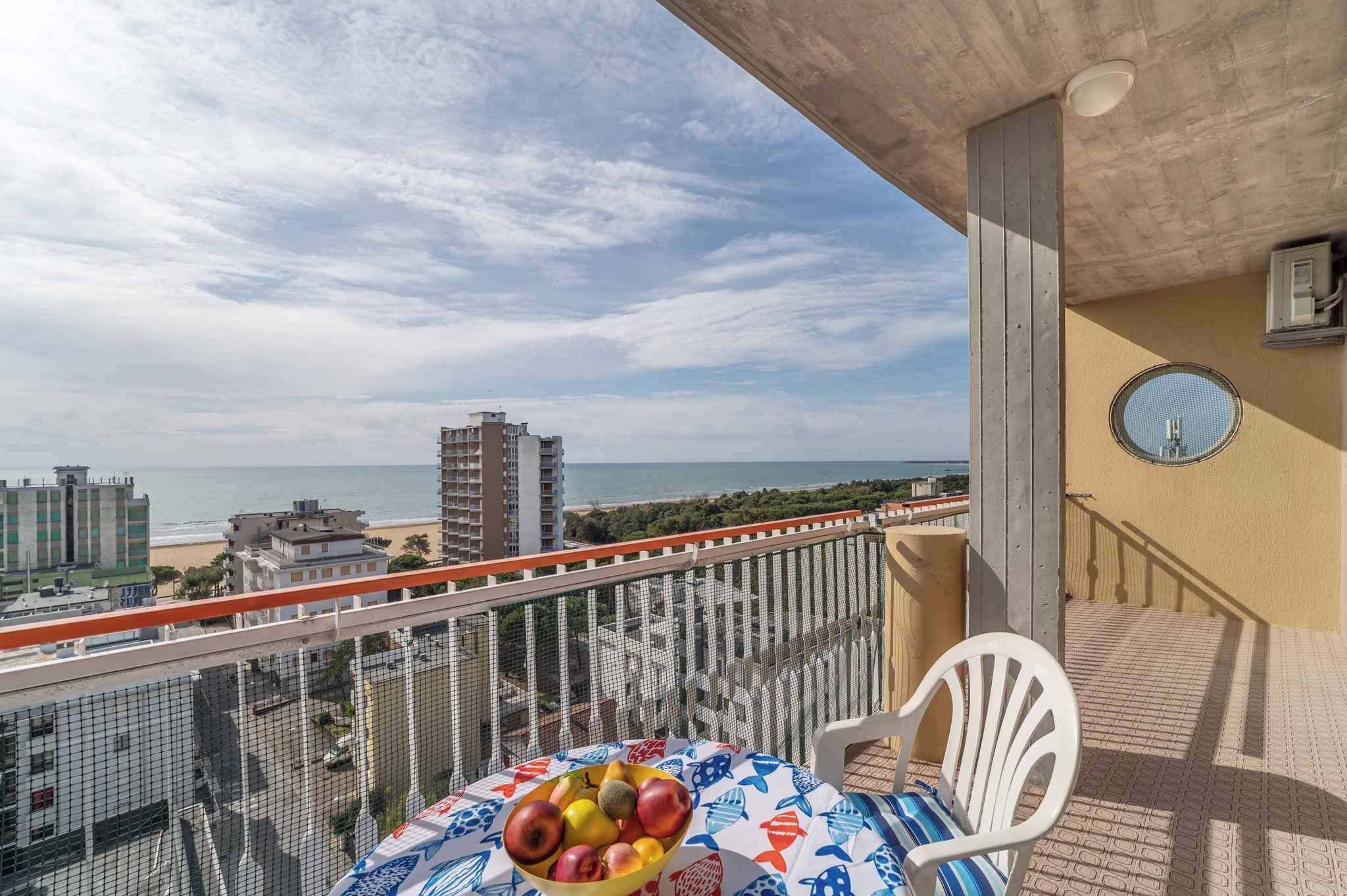 A balcony with a table holding fruit, a chair, and a view of the beach and buildings.
