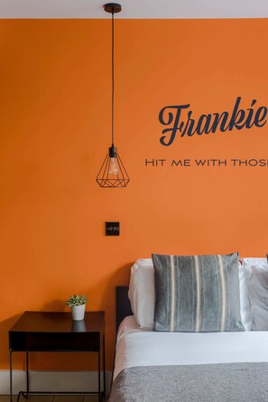 Une pièce avec un mur orange, un lit avec des oreillers, une petite table avec une plante et un luminaire suspendu.