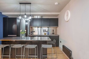 Une cuisine moderne dotée d’appareils électroménagers en acier inoxydable, d’un îlot central avec des tabourets de bar, d’un éclairage suspendu et d’une horloge murale.