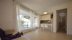 A modern living room with a sofa, a dining area with chairs, a kitchen with a TV, and a balcony with curtains.