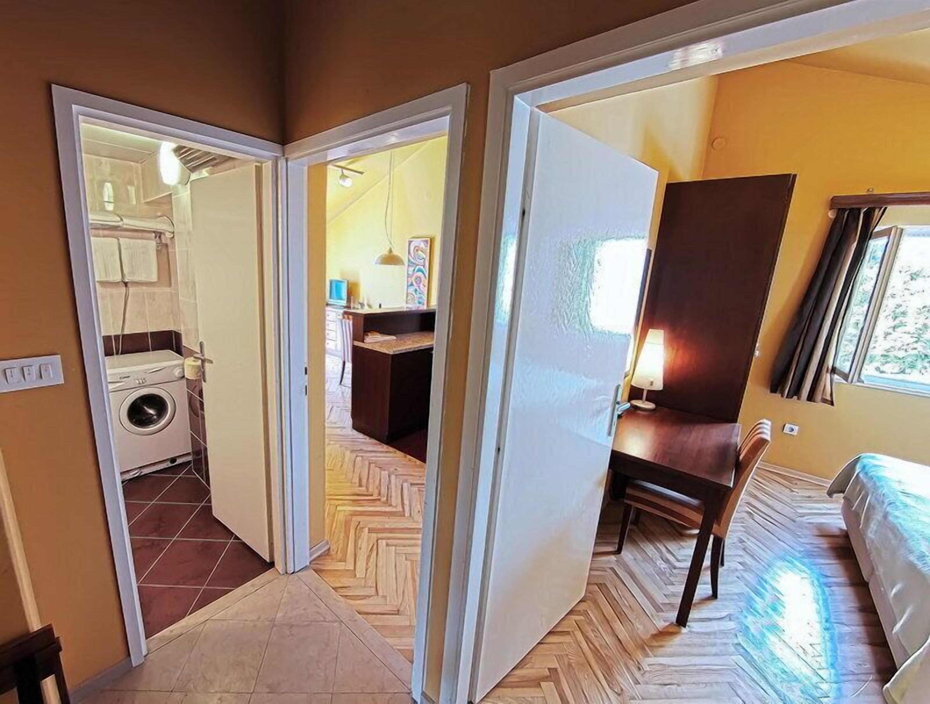 Een kamer met een bed, een bureau en een stoel. Er is een raam met gordijnen en een deur naar een andere kamer.