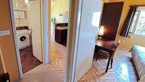 A room with a bed, a desk, and a chair. There is a window with curtains and a door leading to another room.