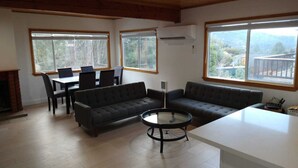 A spacious living room with a dark sofa, a round coffee table, and a dining area with chairs and a table. Large windows provide natural light and views of the outdoors. - Mt. Pinos adjacent Mountain Cabin (Frazier Park)