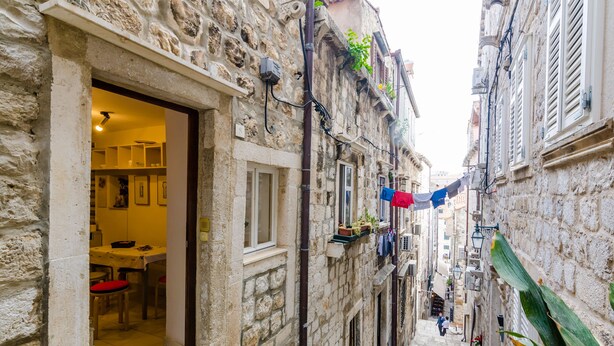 A narrow street with stone buildings, a visible dining area with red chairs, and hanging laundry.
