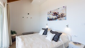 Une chambre à coucher avec un grand lit, des tables de chevet, un banc, une œuvre d’art accrochée au mur et une fenêtre avec des rideaux.