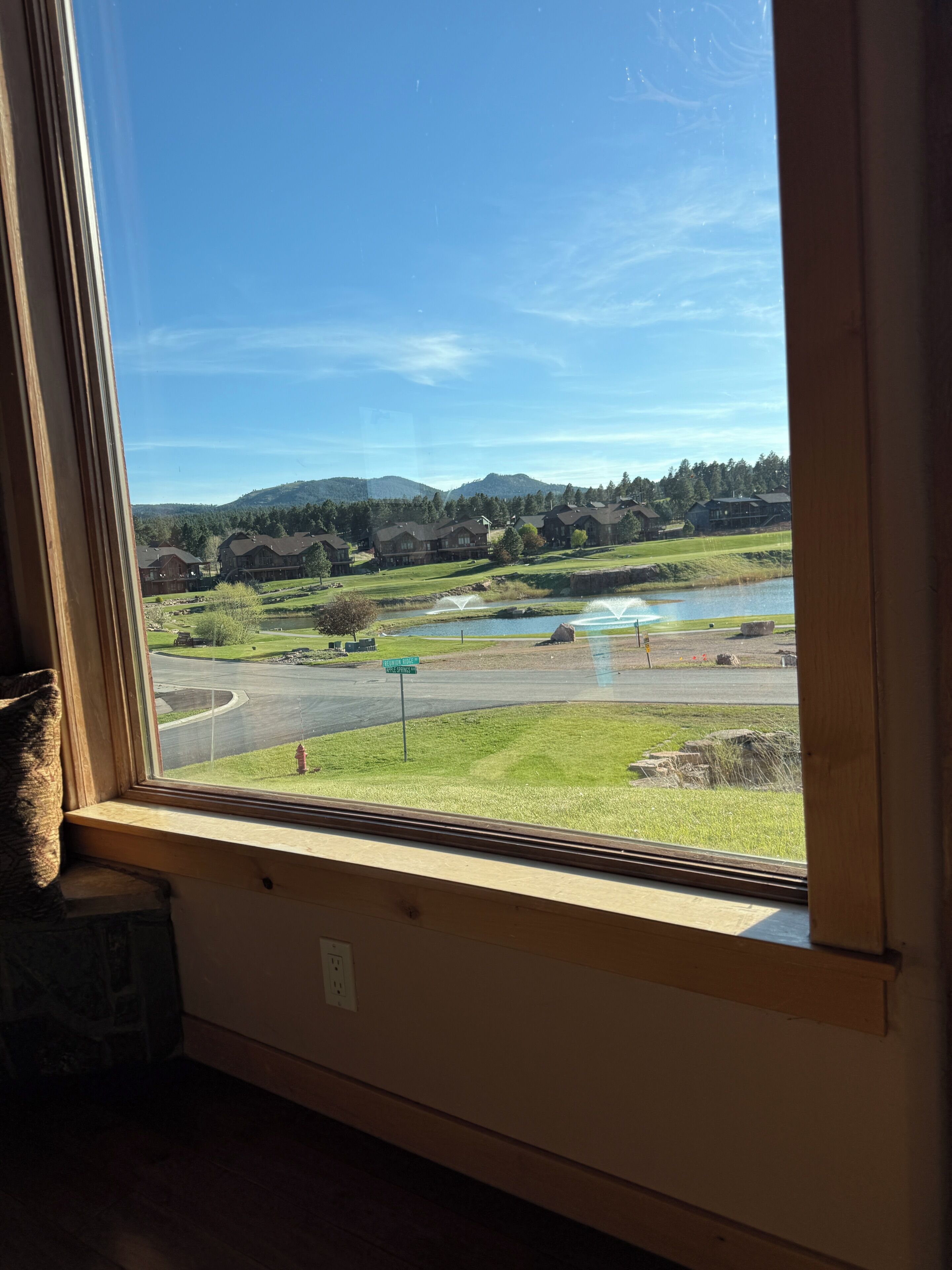 Une vue à travers la fenêtre d’un bâtiment dont le rez-de-chaussée est en pierre, le plancher en bois, et qui offre un panorama sur un terrain de golf, un lac et des collines lointaines.