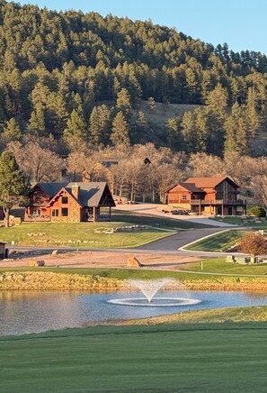 A lodge with a stone exterior, a pond with a fountain, and a forested hill in the background.
