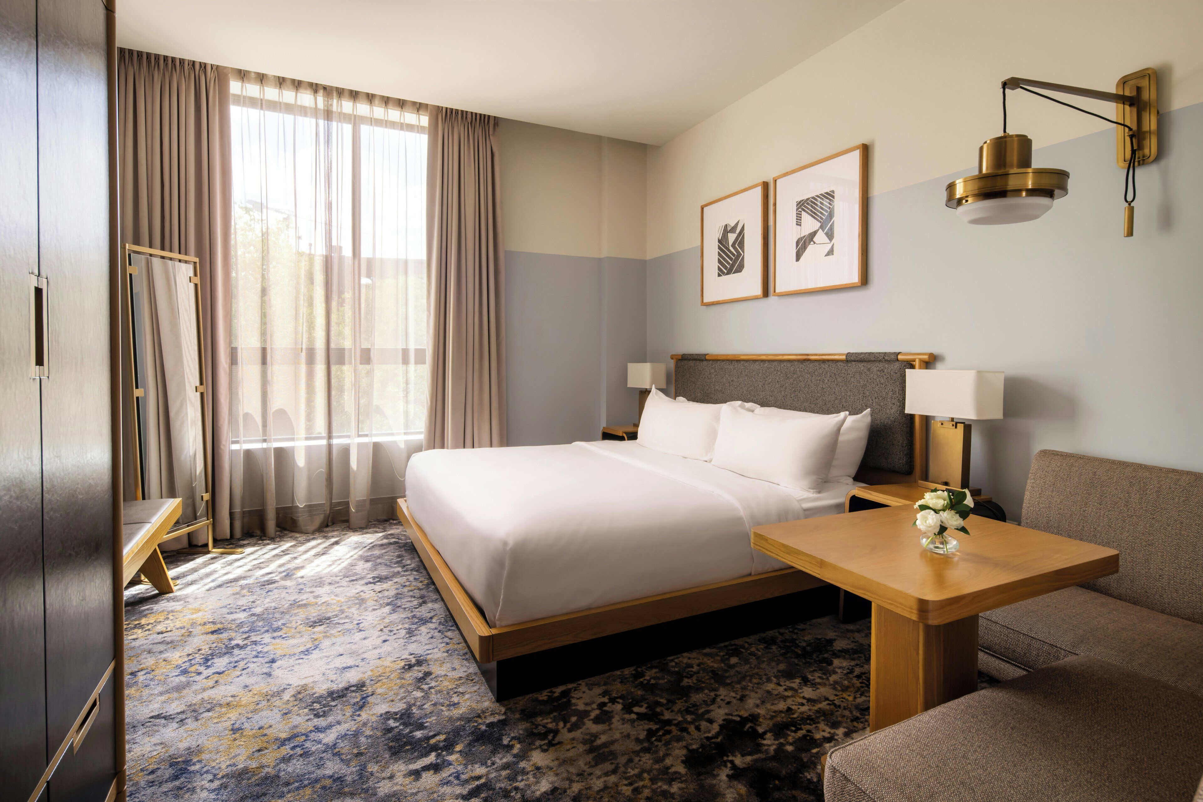 A hotel room with a bed, a bedside table, a lamp, a sofa, a mirror, and a window with curtains.