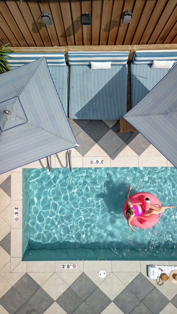 A swimming pool with a person on a pink float, tiled flooring, and umbrellas.