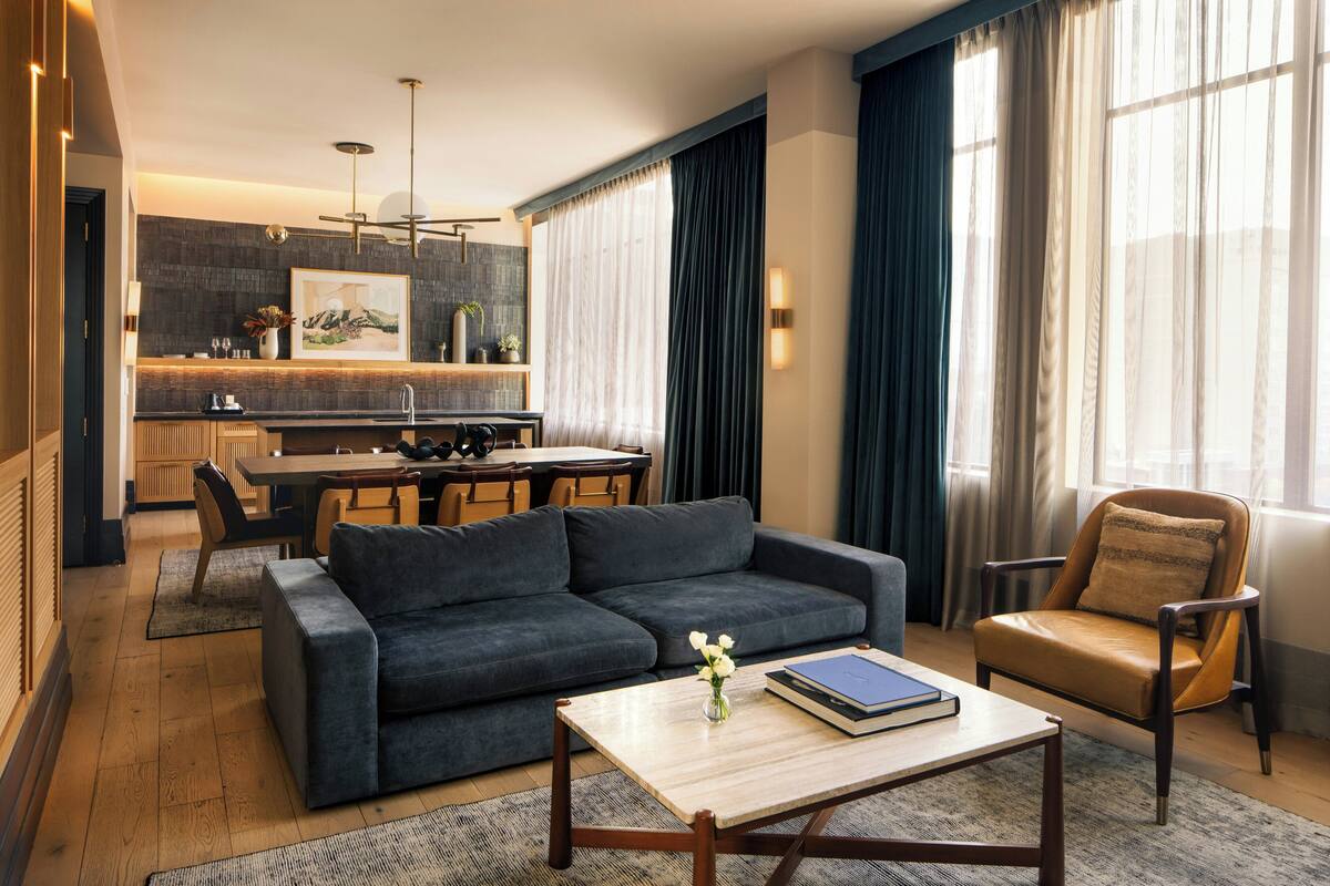 A modern living room with a sofa, armchair, and coffee table, a dining area with a table and chairs, and a kitchen with a countertop and pendant lighting.