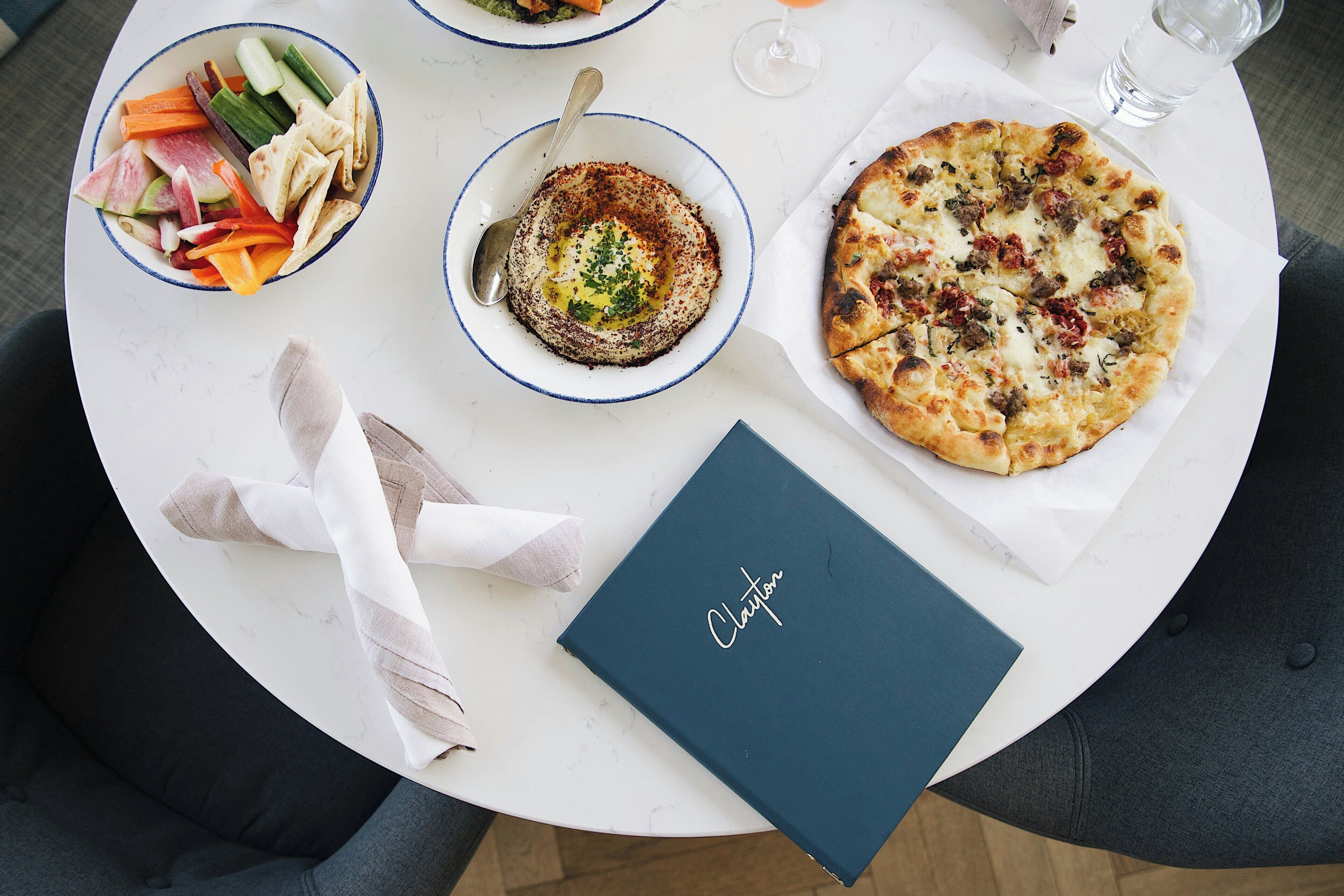 A round dining table with a blue notebook, a bowl of hummus, a plate of vegetables, a bowl of soup, a pizza, and a glass of water.