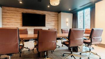 A conference room with a large table, six leather chairs, a flat-screen TV, and a window with curtains.