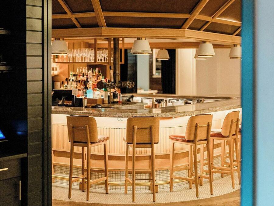 Una zona de bar con sillas de madera y una barra, suelo de baldosas e iluminación colgante.