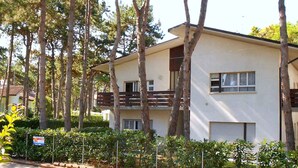 A two-story building with a balcony, surrounded by tall trees and greenery. - Holiday apartment with air conditioning (Lignano)