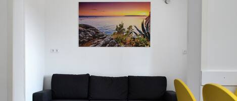 Un salón moderno con un sofá seccional gris oscuro, una butaca amarilla y un cuadro de una puesta de sol sobre un paisaje rocoso en la pared.