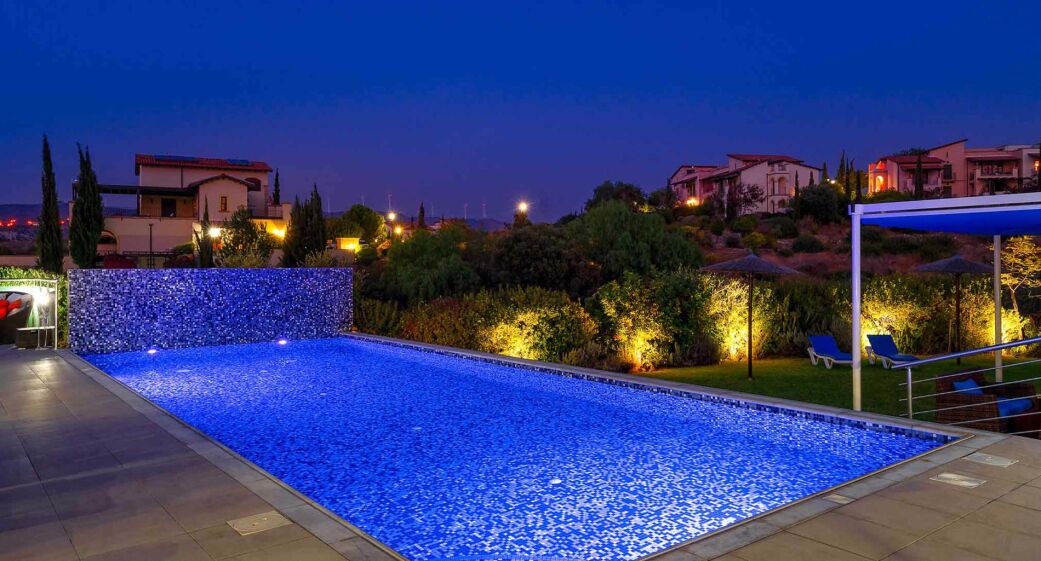 Une piscine dont le bord est revêtu de carrelage, entourée d’un jardin aménagé et dotée d’un espace détente couvert avec des parasols.