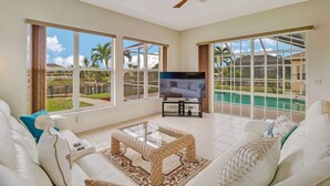 A spacious living room with a large TV, comfortable seating, and large windows offering views of a swimming pool and greenery. - 1226 SE 20th ST (Cape Coral)
