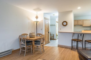 Un comedor con una mesa y sillas de madera, una lámpara colgante, y una zona de cocina con armarios de madera y un reloj en la pared.