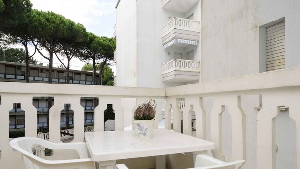A white outdoor seating area with a table and chairs, a potted plant, and a balcony with white railings and a view of trees and a building. - Holiday apartment with air conditioning and pool (Lignano)