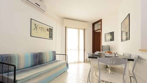 A living room with a sofa, a dining table with chairs, and a kitchen area. - Holiday apartment with air conditioning and pool (Lignano)