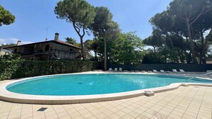 A swimming pool with a tiled deck, surrounded by trees and a building with scaffolding. - Holiday apartment with air conditioning and pool (Lignano)
